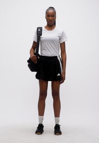 Γυναίκα που στέκεται, φοράει λευκό t-shirt της Adidas, μαύρη φούστα της Adidas, μαύρα αθλητικά παπούτσια, λευκές κάλτσες και κρατάει μαύρη τσάντα ώμου.