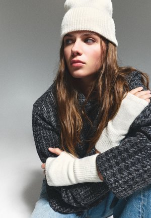 Grijs gebreid oversized sweater met ribgebreide manchetten, gecombineerd met een crèmekleurige beanie. Model toont een ontspannen houding voor een neutrale achtergrond.