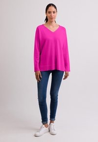 Fuchsie oversized svetr s výstřihem do V, žebrovanými manžetami a lemem. Nošený přes modré skinny džíny a bílé tenisky. Hladká textura.