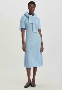 Lichtblauwe gebreide jurk met korte mouwen en een capuchon sjaalkenmerk. Midi-lengte, slank aansluitend ontwerp en structuurstof. Zwarte schoenen.