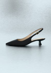 Chaussure slingback noire à bout pointu, petit talon et boucle dorée, présentée de côté sur une surface gris clair.