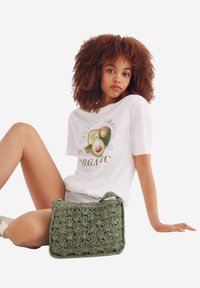 Borsa a uncinetto verde con motivo testurizzato e manico piccolo, posizionata accanto a un modello in t-shirt bianca con un design grafico di frutta.