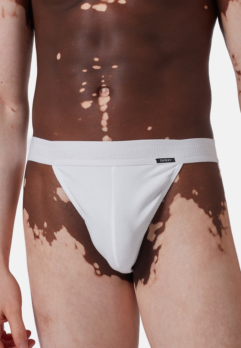 Tanga in cotone bianco con una morbida cintura elastica, decorata con un'etichetta del marchio sulla parte anteriore, indossata da una persona con variazioni della pelle.