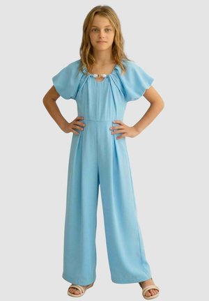 Ragazza giovane in piedi con le mani sui fianchi, indossa una tuta azzurro chiaro a gamba larga e sandali beige su sfondo semplice.