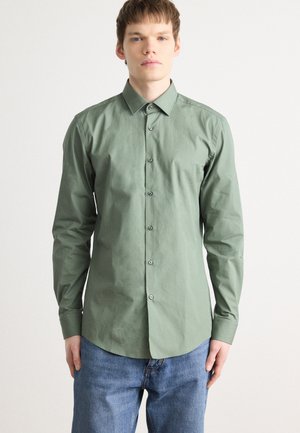 Chemise boutonnée en tissu vert clair, dotée de manches longues, d'un col classique et d'un ourlet droit. Fermée avec des boutons gris.