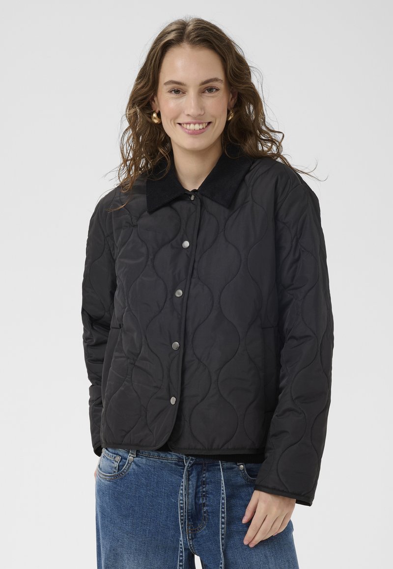 Noor naine laineliste juustega, seljas must tsellutööga jope poppsete kinnitustega ja sinised teksapüksid, naeratab kaamerale valge tausta ees.