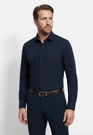 Uomo che indossa una camicia blu navy a maniche lunghe con bottoni, pantaloni abbinati e cintura marrone scuro, guarda di lato.