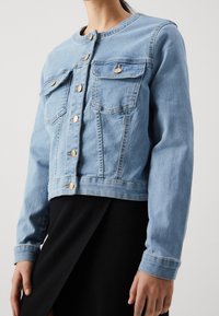 Woman wearing a light blue denim jacket with gold buttons over a black skirt, standing against a plain background.