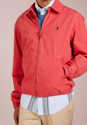 Man draagt een koraalrode jack met rits over een blauwe trui en een gestreept overhemd, met zijn hand in het jackzakje en beige broek.