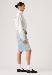 Γυναίκα που στέκεται πλάγια φορώντας γκρι ανοιχτό φούτερ, φούστα από τζιν μέχρι το γόνατο με σκισίματα, λευκές κάλτσες και μαύρα μοκασίνια μπροστά σε απλό φόντο.