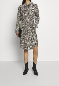 Femme portant une robe longue à manches longues beige et noire à pois avec une ceinture à la taille, associée à des bottines noires et tenant une pochette noire.