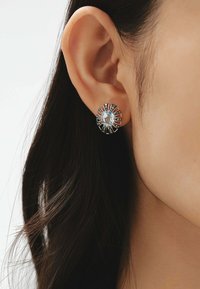 Pendientes de stud en tono plateado que cuentan con una gema azul ovalada en el centro, rodeada de líneas radiales y pequeños acentos claros.