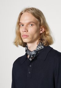Foulard di seta bianco e nero con motivo indossato intorno al collo, caratterizzato da disegni intricati e testo in evidenza, abbinato a una polo blu navy.