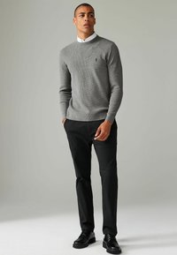 Pull en tricot gris à col rond avec des poignets et un ourlet en côtes, porté sur une chemise blanche à col. Associé à un pantalon noir et des chaussures noires.
