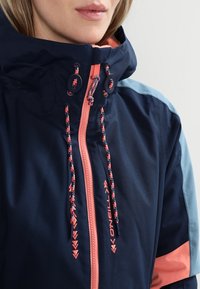 Marineblaue Jacke mit hellblauen Schulterakzenten, hoher Kragen, Reißverschluss vorne und bunten Kordeln mit einem Logo in Pink.