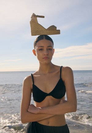 Joven mujer con top de bikini negro está de pie junto al mar con los brazos cruzados, equilibrando una sandalia de cuña sobre su cabeza contra un cielo despejado.