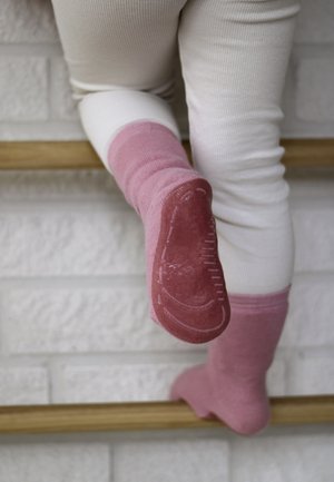 Kind trägt weiße Hose und rosa rutschfeste Socken und klettert an Holzstufen vor einer weißen Ziegelwand.