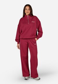 Vínová zipová mikina s vysokým golierom a zodpovedajúcimi širokými nohavicami, vyrobená z mäkkej tkaniny s malým logom na hrudi a nohaviciach.