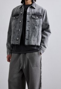 Šedá džínová bunda se dvěma kapsami na prsou, krátkým střihem a roztřepeným lemem, kombinovaná se šedými tepláky s bočními kapsami a stahovací šňůrkou.