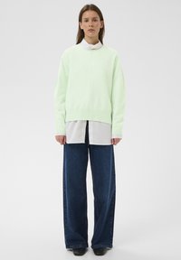 Naine seisab, seljas heleroheline kampsun valge särke peal, kandmas laia säärega tumesiniseid pükse ja musti kingi, taustaks tavaline taust.