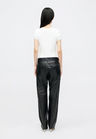 Γυναίκα με μακριά μαύρα μαλλιά, φορώντας ένα εφαρμοστό λευκό μπλουζάκι κοντό μανίκι, μαύρο δερμάτινο παντελόνι ψηλής μέσης και μαύρες ψηλοτάκουνες γόβες, όρθια.