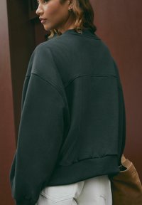 Felpa nera oversize con spalle scese, polsini a costine e una texture elegante. Abbinata a pantaloni chiari e a una borsa marrone.