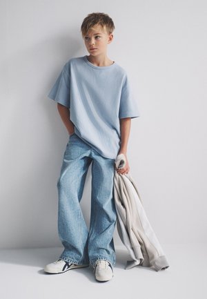 Dječak u širokoj svijetloplavoj majici i širokim trapericama, držeći svijetlosivu jaknu, stoji uz običan bijeli zid.