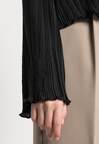 Blusa nera a maniche lunghe plissettata con orlo arricciato e tessuto testurizzato. Indossata sopra un capo beige, che mette in mostra elementi di design a strati.