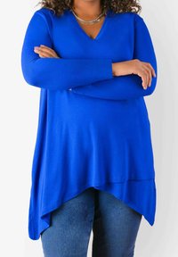 Kungsblå långärmad topp med V-hals, med asymmetrisk fåll och mjuk textur, tillsammans med denimjeans.