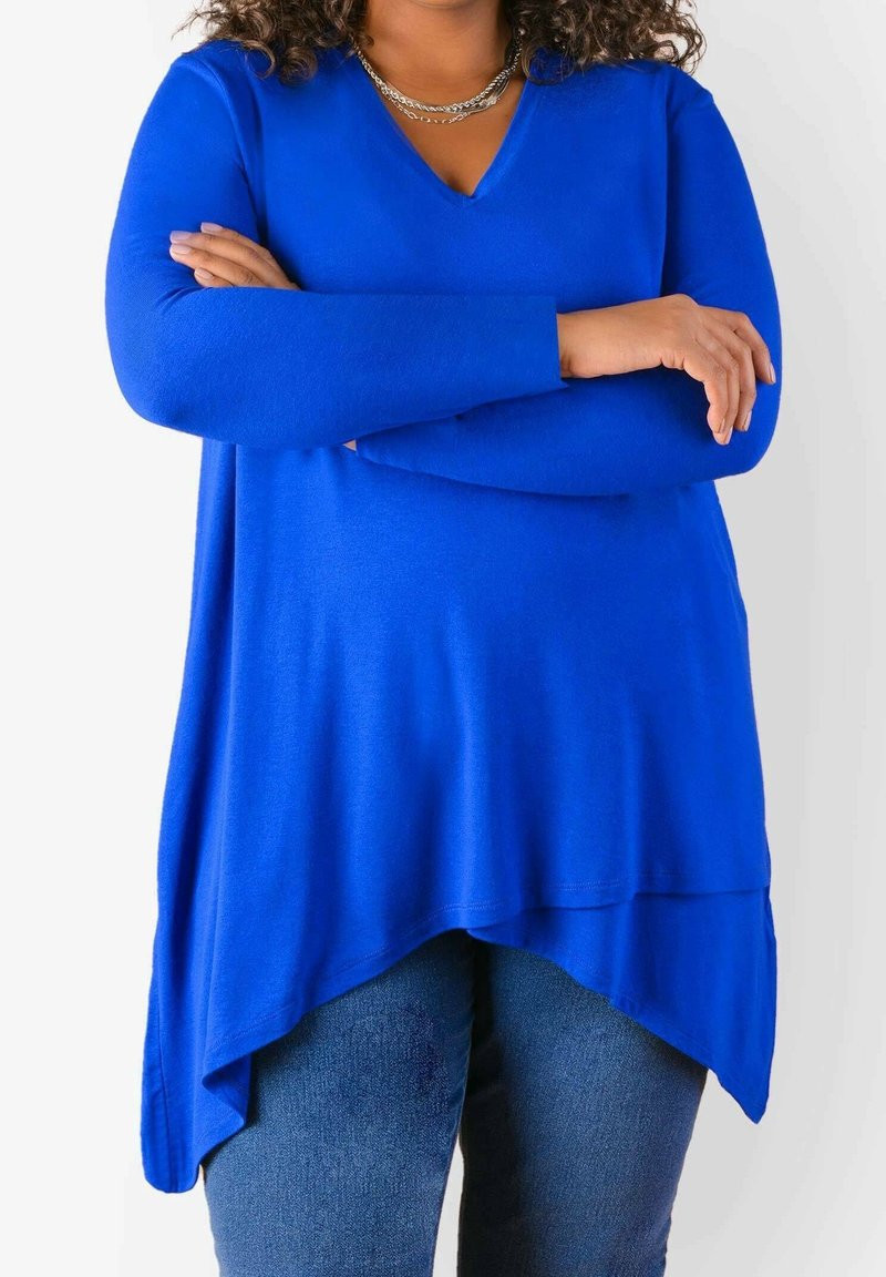 Kungsblå långärmad topp med V-hals, med asymmetrisk fåll och mjuk textur, tillsammans med denimjeans.