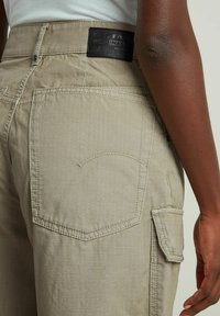 Pantalon cargo beige en tissu texturé, avec un détail de poche cousue et une étiquette noire à la taille. Poche utilitaire latérale incluse.