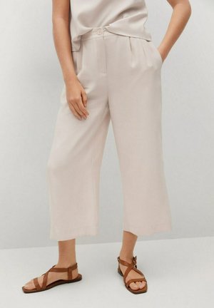 Femme portant un pantalon beige à jambes larges et des sandales plates marron, se tenant avec une main dans sa poche sur un fond neutre.