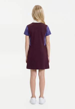Kind met lang blond haar draagt een paarse jurk met blauw gemêleerde korte mouwen en een witte kraag, staat met de rug naar de camera in witte sneakers.