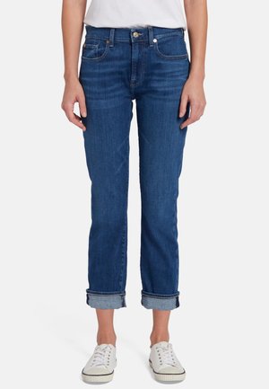 Jeans in denim blu con vestibilità dritta, orli risvoltati, leggera sbiaditura e design classico a cinque tasche, indossati con sneakers bianche.