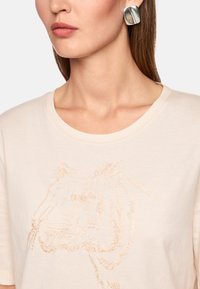 Beige cotton t-shirt featuring a tiger embroidered in gold thread. Short sleeves and a round neckline. Large silver geometric earrings.