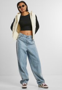 Gilet trapuntato crema sopra una maglietta nera a maniche lunghe, abbinato a jeans larghi azzurri e sneakers marroni con dettagli rosa.