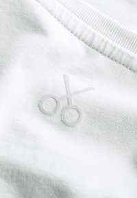 Witte katoenen stof met een zachte textuur, voorzien van een subtiele grijze schaarlogo geborduurd in het midden. Ronde halslijn zichtbaar.