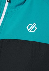 Turquoise en zwarte jas met een gladde textuur, ritssluiting en een wit geometrisch logo op het turquoise gedeelte.