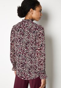 Donna con i capelli ricci raccolti in una coda di cavallo, che indossa una blusa a maniche lunghe con un motivo viola, nero e bianco e pantaloni bordeaux, in piedi di lato.