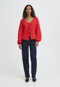 Cardigan rosso lavorato a maglia con scollo a V, maniche lunghe e chiusura con laccio sul davanti, abbinato a jeans blu scuro a gamba dritta e scarpe nere con punta affilata.
