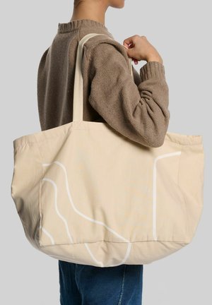 Person trägt einen braunen Pullover und eine große beige Tragetasche mit weißen abstrakten Linien auf der Schulter vor einem grauen Hintergrund.
