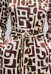 Abito marrone e crema con motivo a fantasia, chiusura con bottoni davanti, cintura annodata in vita e maniche lunghe; presenta forme geometriche e tessuto dalla texture liscia.