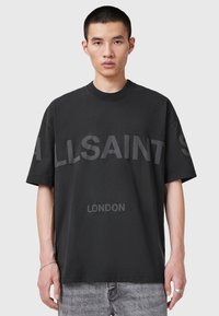 Tânăr cu păr scurt poartă un tricou oversized negru cu textul "ALLSAINTS" și "LONDON", asortat cu blugi gri deschis.