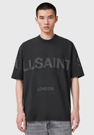 Junger Mann mit kurzen Haaren trägt ein schwarzes Oversize-T-Shirt mit den Aufdrucken "ALLSAINTS" und "LONDON", kombiniert mit hellgrauen Jeans.
