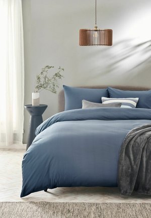 Bett mit blauer Bettwäsche und Kissen, grauer Decke, hölzerner Pendelleuchte und Beistelltisch mit Pflanze in neutralem Schlafzimmer.