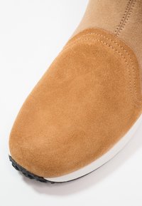 Bota de ante marrón claro con una textura suave, punta redondeada y una entresuela blanca en contraste. Presenta costuras reforzadas a lo largo del borde. Suela negra.