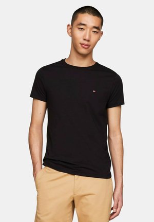 Giovane uomo con capelli corti che indossa una maglietta nera semplice e pantaloni beige, in piedi con una mano in tasca contro uno sfondo bianco.