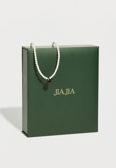 Πράσινο κουτί JIAJIA με ένα λευκό μαργαριταρένιο κολιέ να κρέμεται πάνω του, με ένα σκούρο μενταγιόν σε σχήμα δάκρυ.