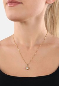 Donna che indossa una sottile collana d'oro con un pendente rotondo che presenta una pietra preziosa trasparente, mostrata dal collo fino alla parte superiore del petto in una maglietta nera.
