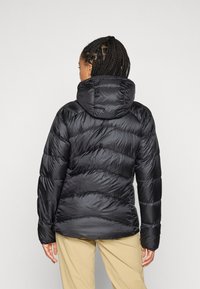 Persoon met gevlochten haar, gekleed in een zwarte gewatteerde pufferjack met capuchon en beige broek, staande tegen een effen lichte achtergrond.